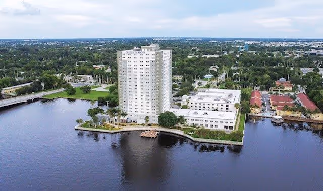 Aerial view of Calusa Harbour, a tall white multi-story building situated on a waterfront with surrounding greenery and other buildings in the background under a cloudy sky.
