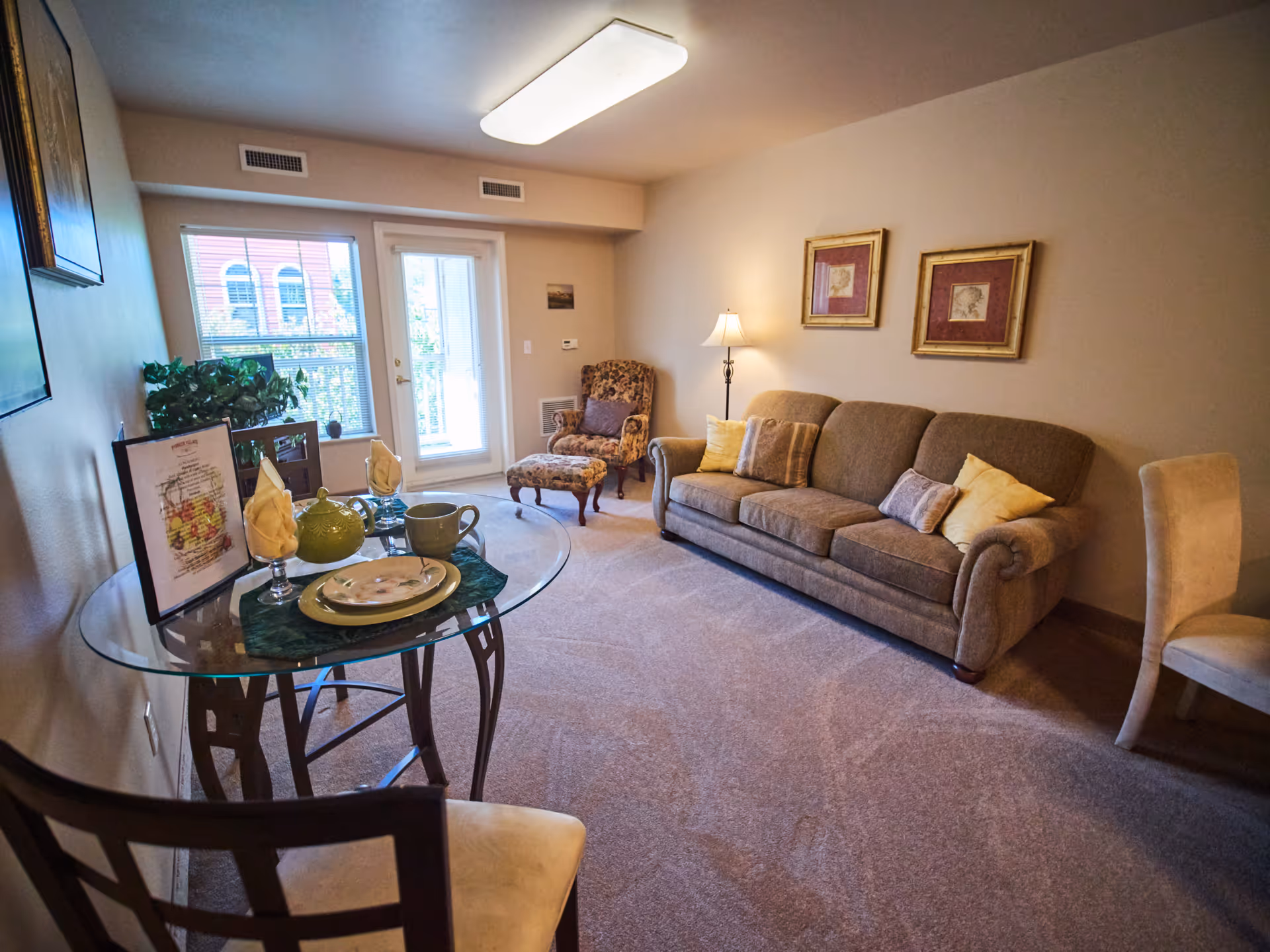 A cozy living room with a beige sofa adorned with multiple cushions, a patterned armchair with an ottoman, a floor lamp, and two framed pictures on the wall. There is a glass-top table set with plates, cups, and napkins, and a chair nearby. A window and a glass door leading to a balcony allow natural light to enter the room.