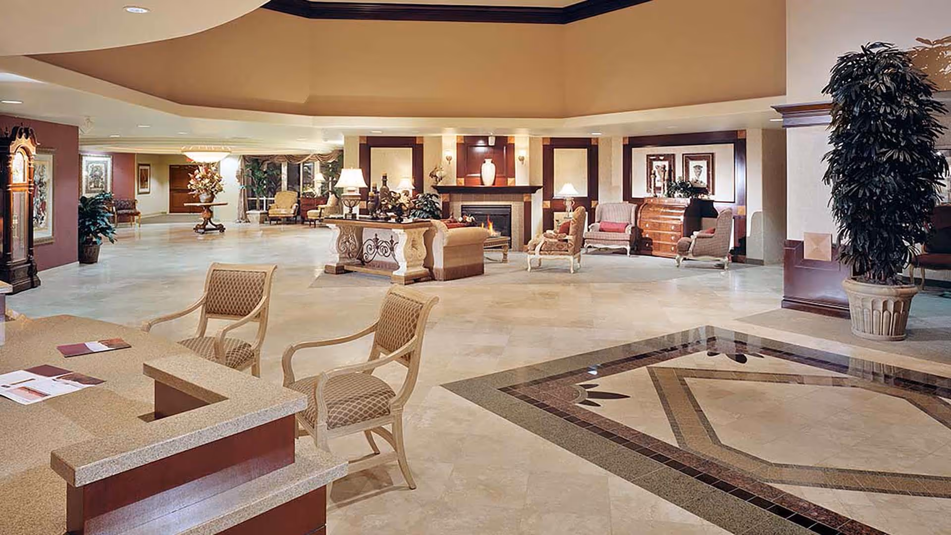 Spacious and elegant senior living facility lobby with a large tiled floor, comfortable seating areas including armchairs and sofas, a fireplace, decorative plants, and a reception desk with chairs.