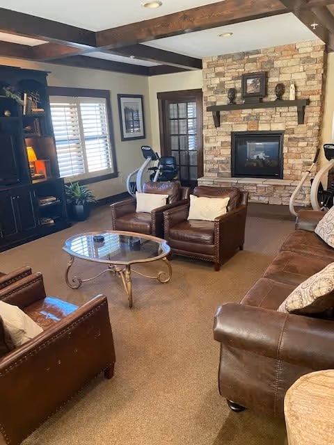 Cozy living room with brown leather chairs and sofa arranged around a glass coffee table. A stone fireplace with a wooden mantle is centered on the far wall, decorated with small sculptures and a framed picture. There is a dark wooden bookshelf on the left side near a window with white blinds. Exercise equipment is visible near the back wall, and the room has exposed wooden ceiling beams.