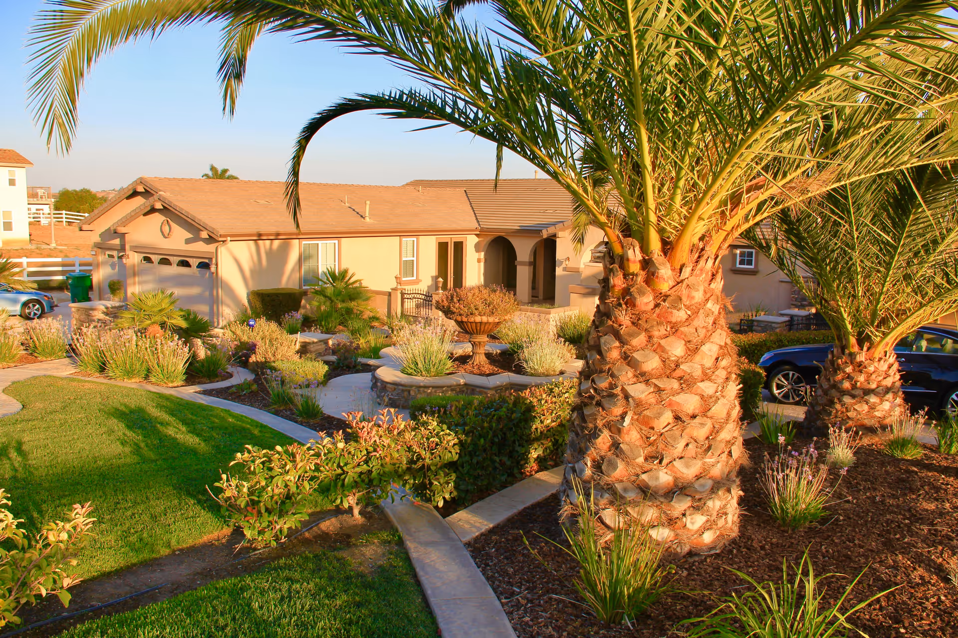 A sunny outdoor view of a single-story beige building with a tiled roof, surrounded by landscaped gardens featuring palm trees, shrubs, and a circular stone planter. A driveway with parked cars is visible on the right side.