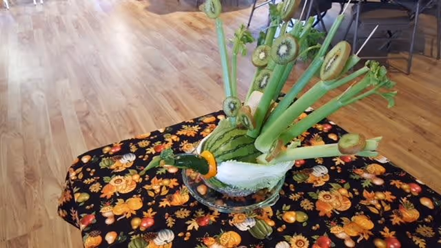 A decorative centerpiece on a table featuring a watermelon carved and arranged with celery stalks and sliced kiwi pieces, placed on a black tablecloth with a colorful autumn-themed pattern. The background shows a wooden floor and some chairs and tables.