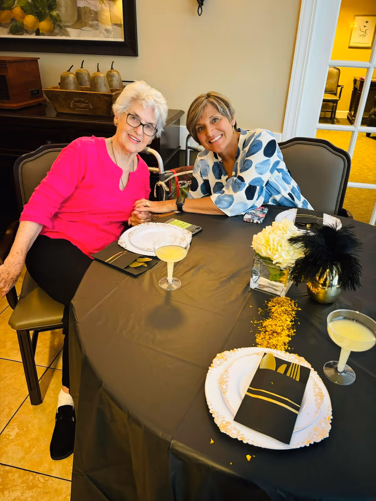 Two women sitting at a dining table in a well-lit room. One woman with white hair and glasses is wearing a bright pink top, and the other woman with short hair is wearing a white blouse with blue polka dots. They are smiling and holding hands across the table, which is covered with a black tablecloth and set with decorative plates, black napkins with gold cutlery, two cocktail glasses with a light-colored drink, and a centerpiece with white flowers and black feathers.