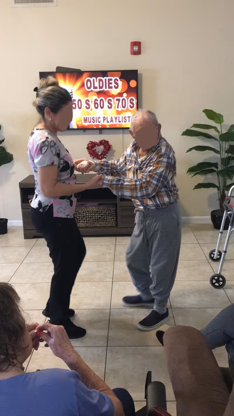 An elderly man and a woman holding hands and dancing in a living room area. A television in the background displays a colorful screen with the text 'OLDIES 50'S 60'S 70'S MUSIC PLAYLIST'. There are plants on either side of the TV stand, and two other elderly individuals are seated nearby, one in a wheelchair.