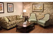 A cozy living room with a floral patterned sofa, two light green upholstered armchairs, a dark wooden coffee table with decorative items, a floor lamp, and framed artwork on a stone accent wall.