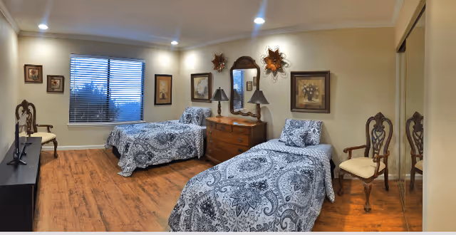A senior care facility bedroom with two single beds covered in matching blue and white patterned bedspreads. The room has wooden flooring, a wooden dresser with two lamps and a mirror above it, framed artwork on the walls, a window with blinds, and two wooden chairs. There is a TV on a stand opposite the beds.