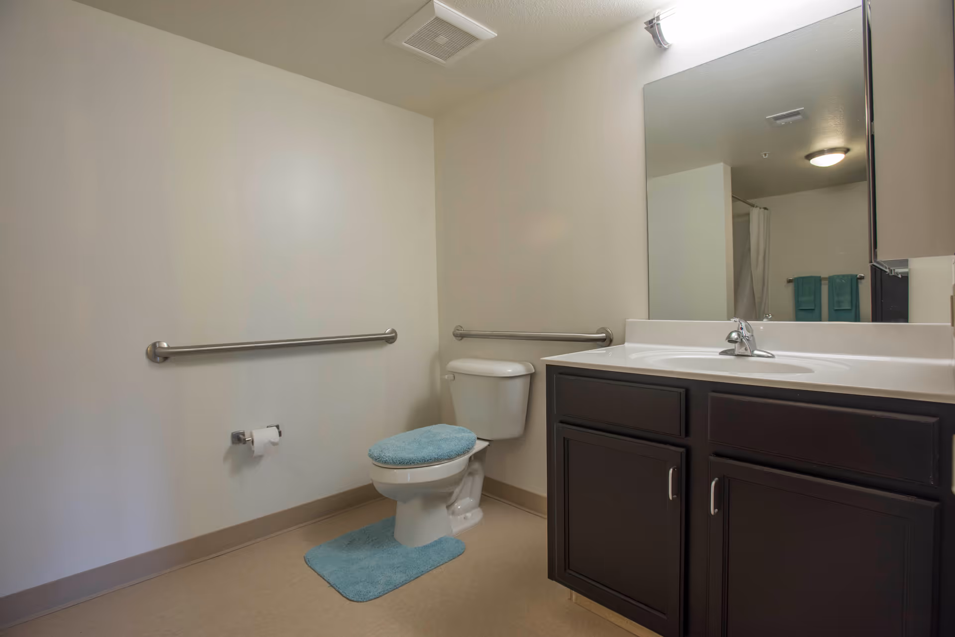 A clean bathroom with beige walls and flooring, featuring a white toilet with a blue cushioned seat cover and matching blue rug. There are stainless steel grab bars on the walls near the toilet. A dark wood vanity with a white countertop and sink is visible, along with a large mirror above it. In the reflection, a shower curtain and two blue towels hanging on a rack can be seen.