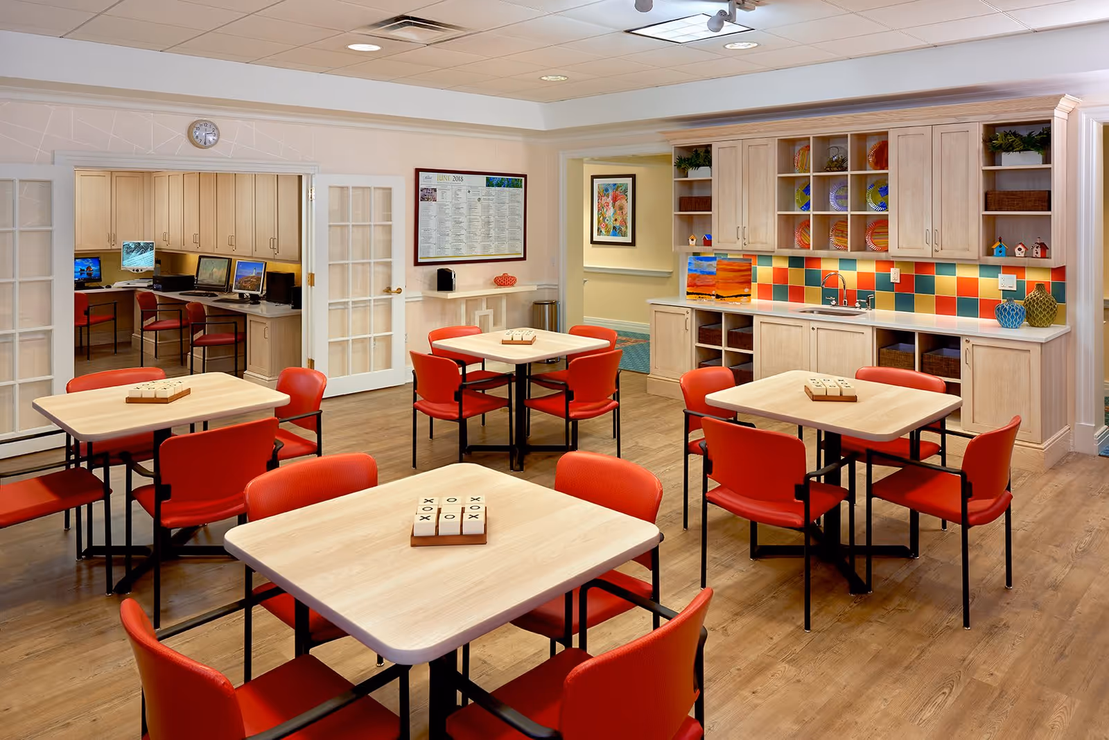 Bright communal dining/activity room with several square tables and red chairs, a kitchenette with a colorful tile backsplash, and a computer station visible through French doors.