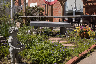 Outdoor garden area at Penacook Place featuring a stone bench engraved with 'In Memory of Kathy Keefe', surrounded by flowers, plants, a small statue of a child holding a basket, and a brick pathway leading to a patio with tables and chairs.