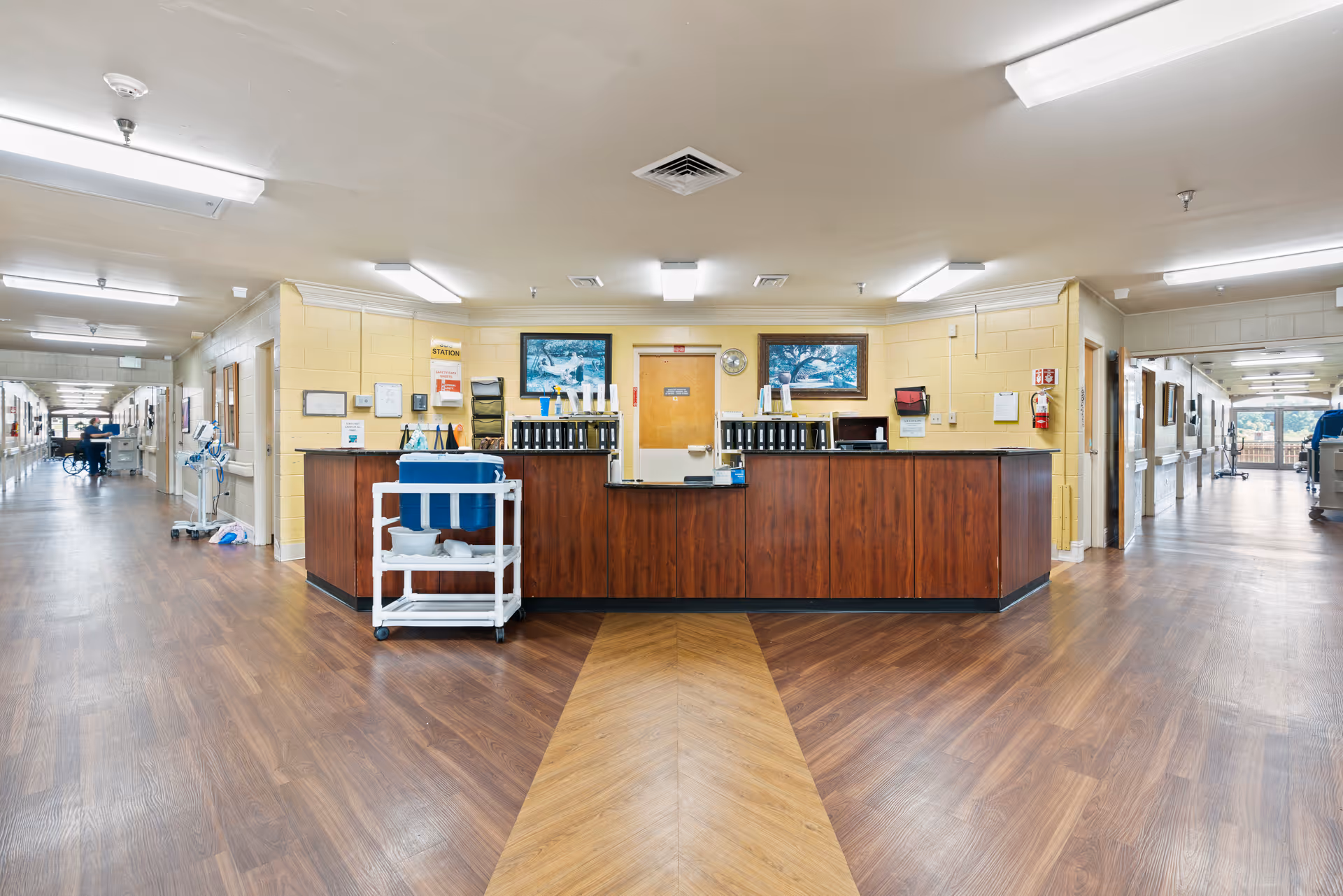A wide hallway in a senior living facility with a central wooden reception desk. The floor has a wood pattern, and the walls are painted light yellow. There are two framed pictures above the desk, a clock, and various office supplies. The hallway extends on both sides with handrails along the walls and medical equipment visible in the distance.