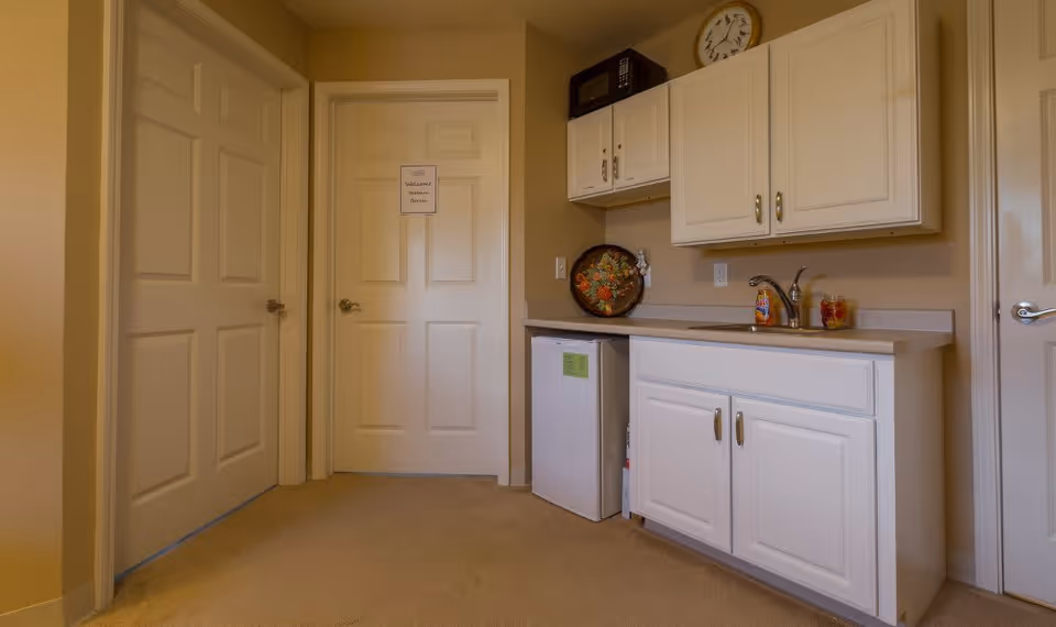 Small kitchenette area with white cabinets, a countertop with a sink, a mini refrigerator, a microwave on top of the upper cabinets, and a decorative tray. There are three closed doors visible, one with a sign that reads 'Welcome Eastern Garden.' The room has beige walls and carpeted floor.