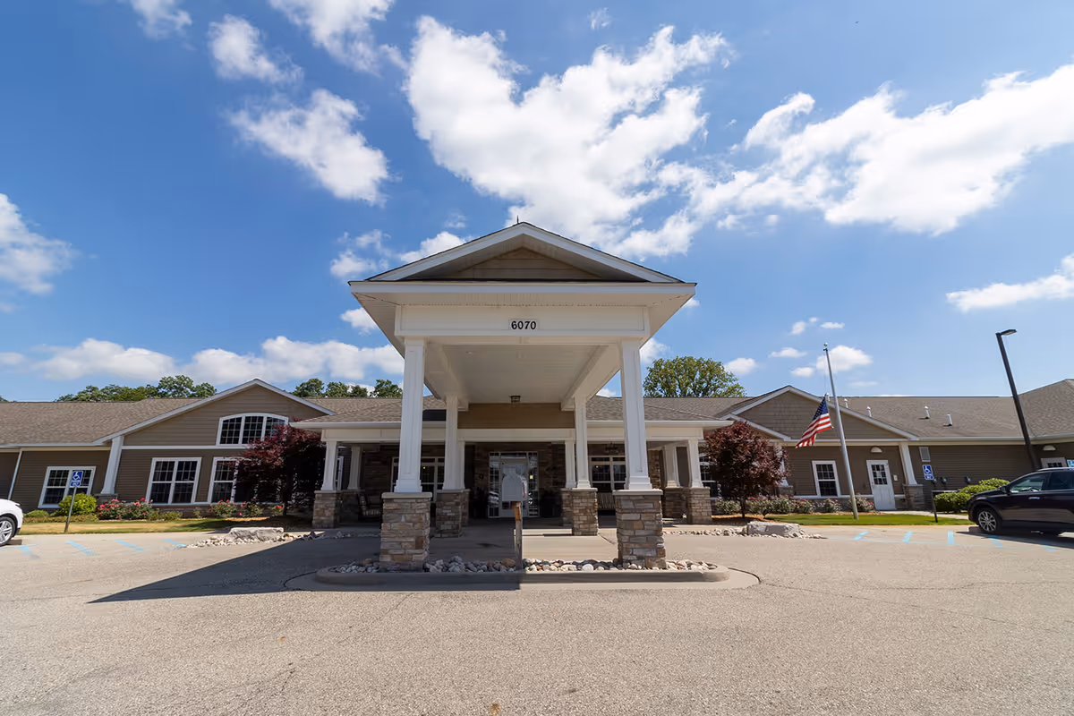 Front entrance of a single-story assisted living facility with a covered portico, stone columns, and an American flag under a blue sky.