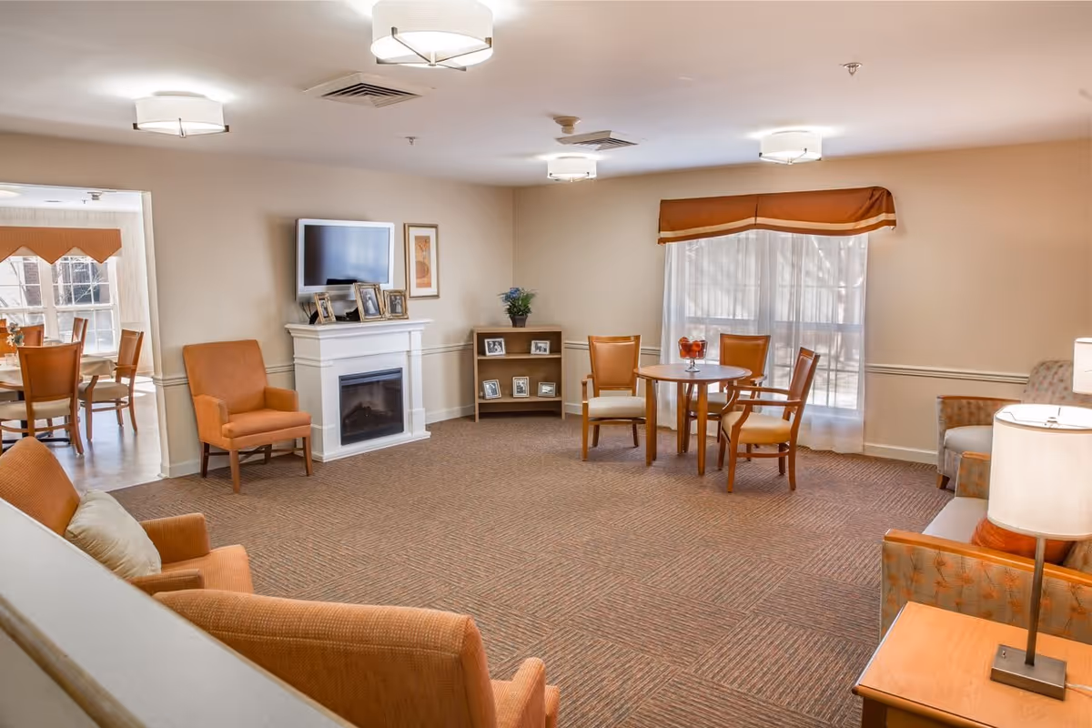 Bright senior living community common room with seating, a fireplace under a wall-mounted TV, and a small round dining table.