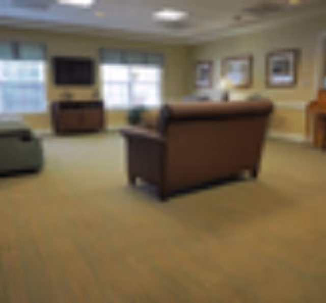 A living room area with a brown sofa facing away from the camera, a television mounted on the wall, windows with blinds, framed pictures on the walls, and a piano in the corner.