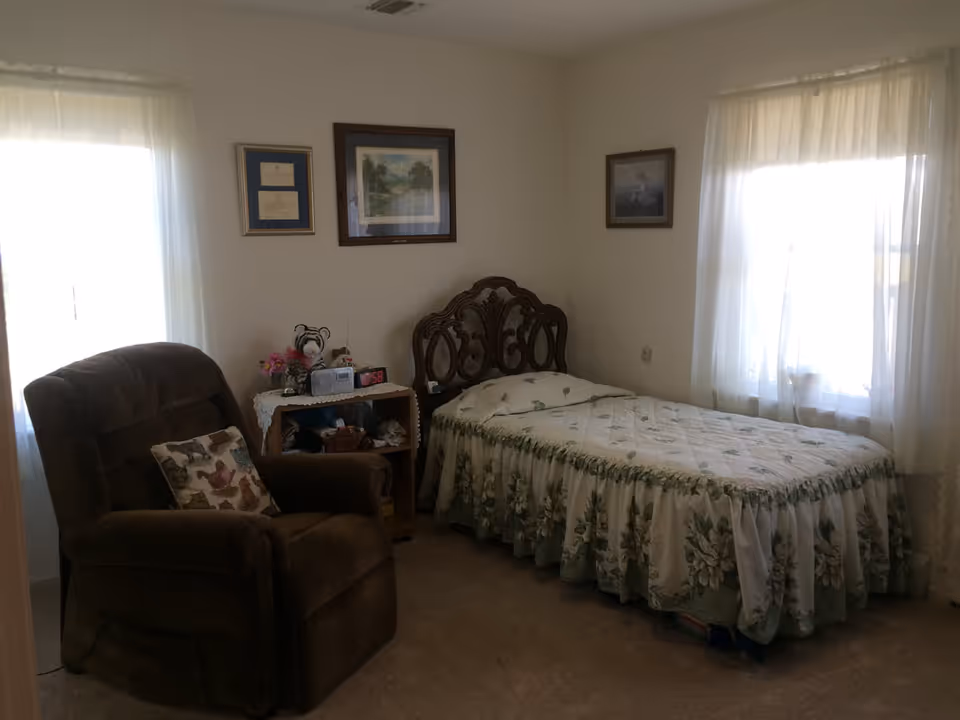 A cozy bedroom with a single bed covered in a floral bedspread, a brown recliner chair with a decorative pillow, a small wooden nightstand with a radio, flowers, and other small items, and three framed pictures hanging on the walls. Two windows with sheer white curtains allow natural light into the room.