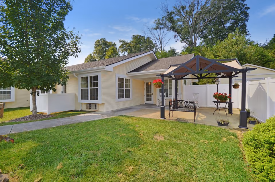 Outdoor patio area of a single-story building with beige siding and white trim, featuring a covered pergola with hanging flower pots, a bench, a table with flowers, and a white fence surrounding the patio. There is a green lawn and trees in the background under a blue sky.