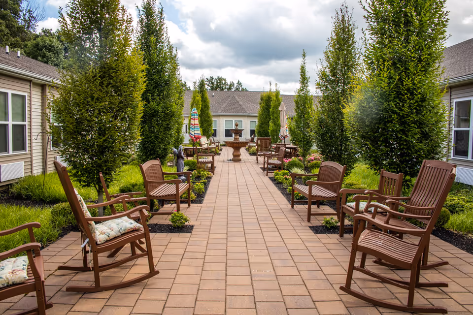 Outdoor patio area at Mullica Gardens Assisted Living with a paved walkway lined by tall green trees and brown wooden rocking chairs with cushions on both sides. In the background, there are more chairs, tables with umbrellas, and a water fountain, surrounded by well-maintained greenery and buildings.