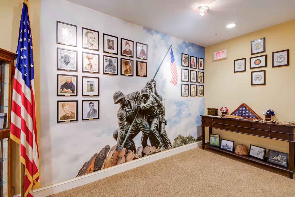 A memorial corner inside a senior living facility featuring a large mural of the iconic Iwo Jima flag raising statue against a sky background. Surrounding the mural are framed photographs of veterans and military personnel. An American flag stands on the left side, and a wooden console table on the right holds a folded flag in a triangular display case, framed pictures, and decorative items. The walls are painted light yellow and beige, and there is an exit sign above the console table.