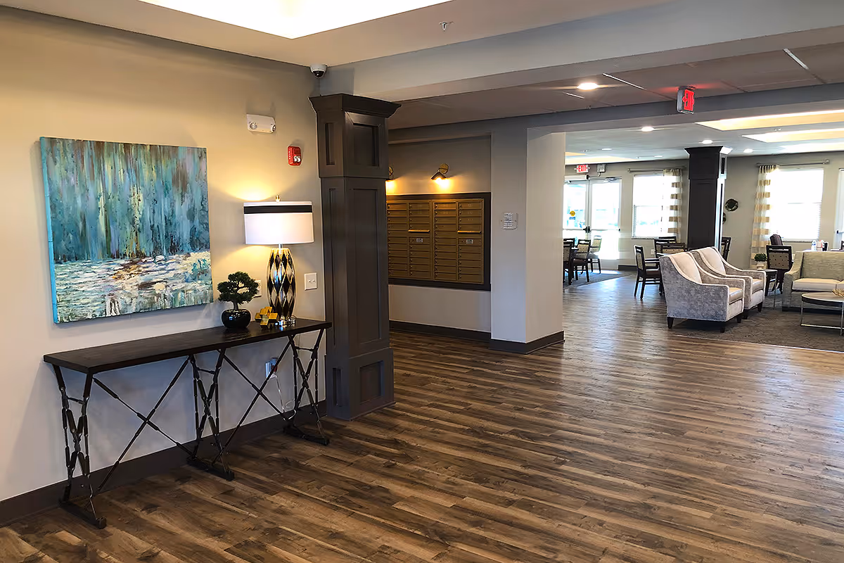Interior view of a senior living facility lobby area with wood flooring, a decorative table with a lamp and small plant, a large abstract painting on the wall, mailboxes mounted on the wall, and a seating area with chairs and sofas in the background near large windows.