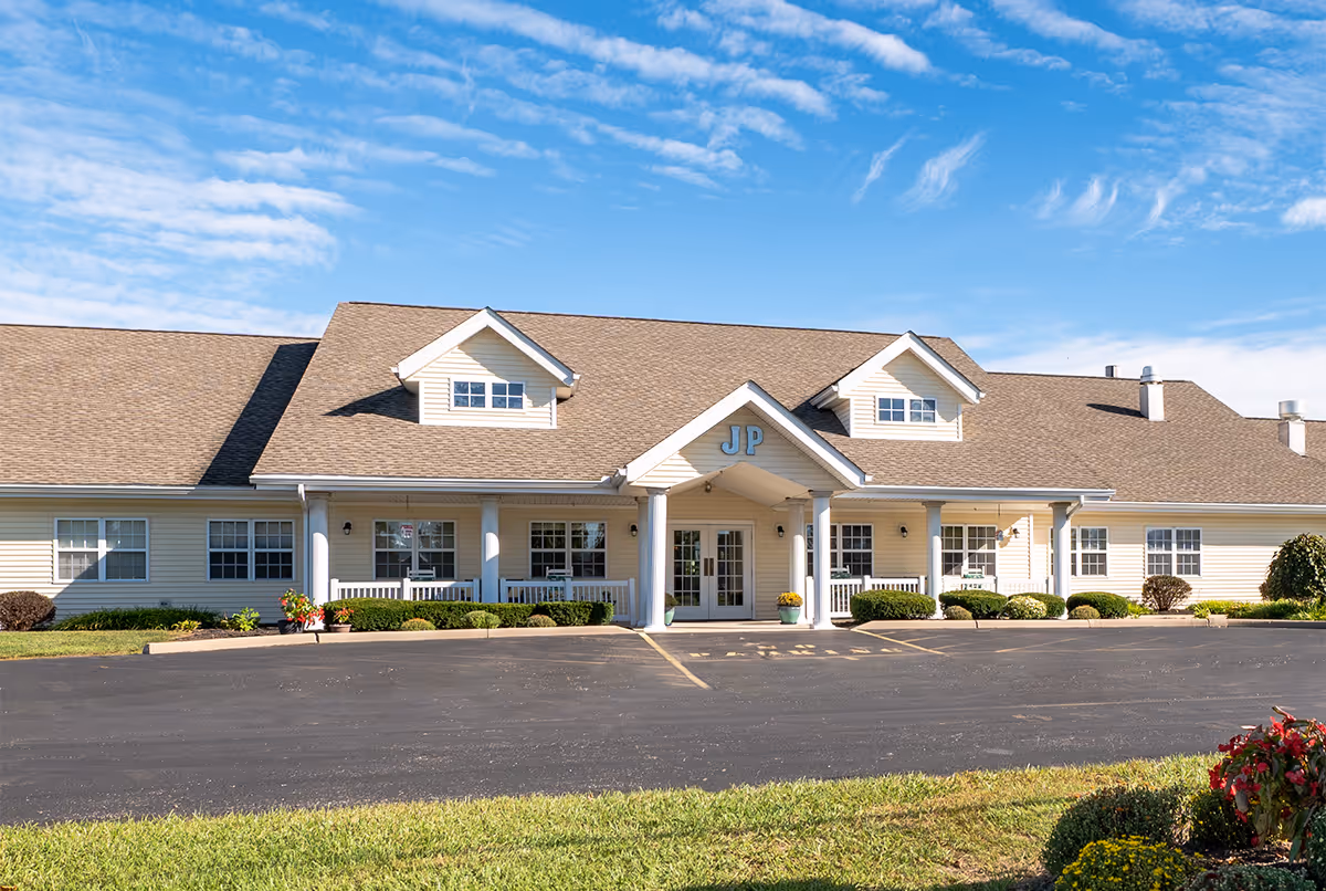 Exterior view of a single-story senior living facility building with beige siding and a brown shingled roof under a blue sky with scattered clouds. The entrance features a covered porch with white columns and the letters 'JP' above the door. There are shrubs and flowers planted along the front of the building and a paved parking area in front.