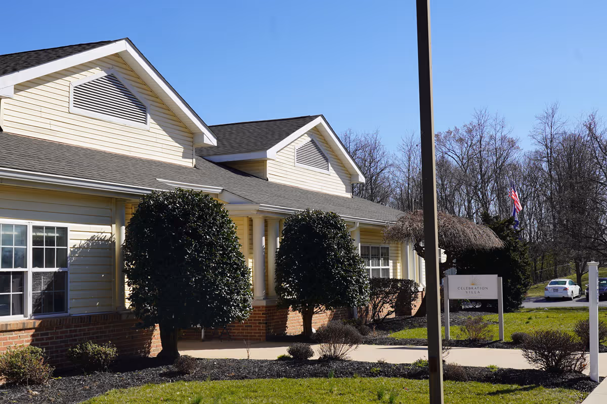 Exterior view of Celebration Villa of Martinsburg, a single-story building with yellow siding and a gray roof. The building is surrounded by neatly trimmed bushes and landscaping, with a sidewalk leading to the entrance. A sign with the facility's name is visible near the entrance, and a few cars are parked in the background under a clear blue sky.