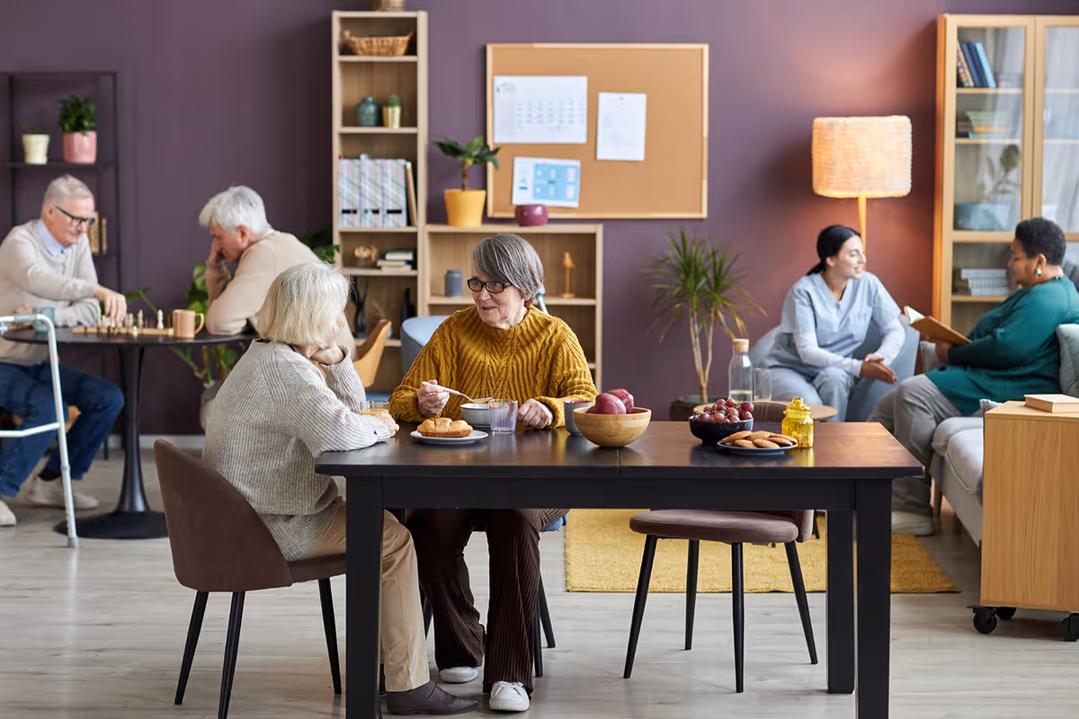 Elderly residents sit at a communal table eating and talking in a cozy senior living common room while others play chess and converse in the background.
