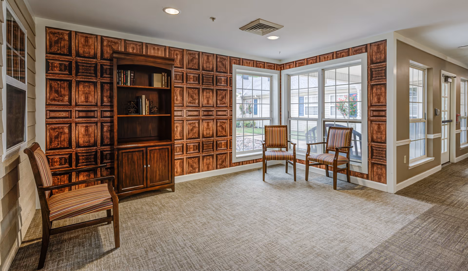 A quiet sitting area in a senior living facility with three wooden chairs featuring striped cushions, a wooden bookshelf with a few books and decorative items, and large windows letting in natural light. The walls have a decorative wooden panel design and the floor is carpeted.