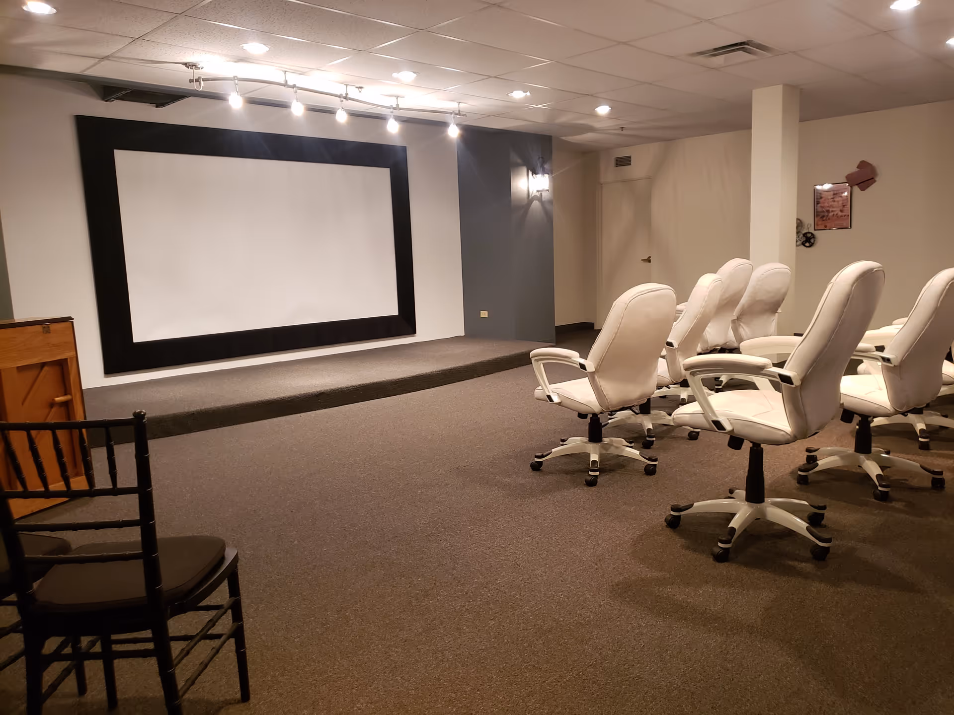 A small theater room with a large white projection screen framed in black on a raised platform. In front of the screen are several white swivel chairs arranged in rows, and a black wooden chair is visible in the foreground. The room has a carpeted floor and a drop ceiling with recessed lighting.