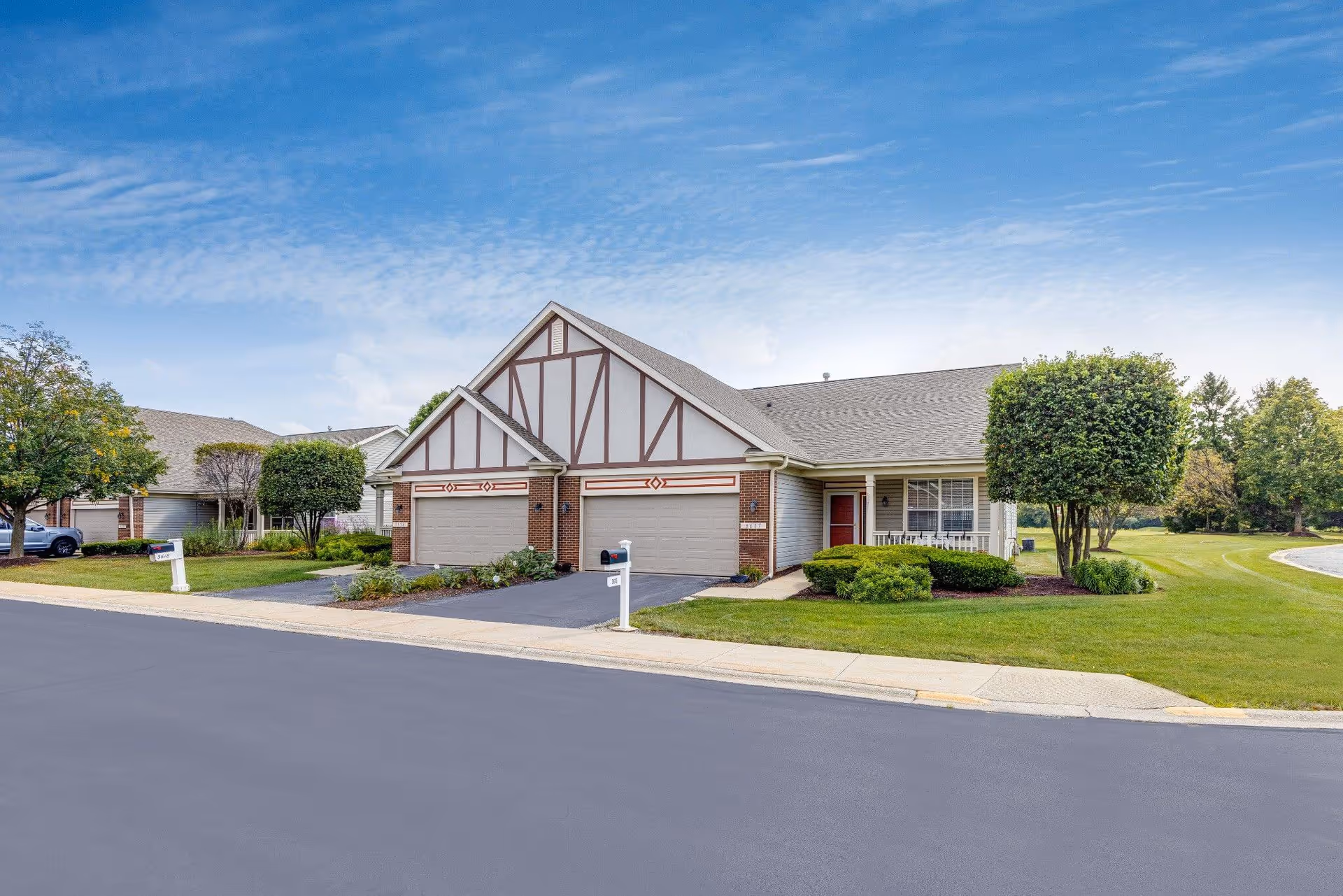 Exterior view of a residential-style building with two garages, a small porch, and well-maintained landscaping including bushes and trees under a partly cloudy sky.