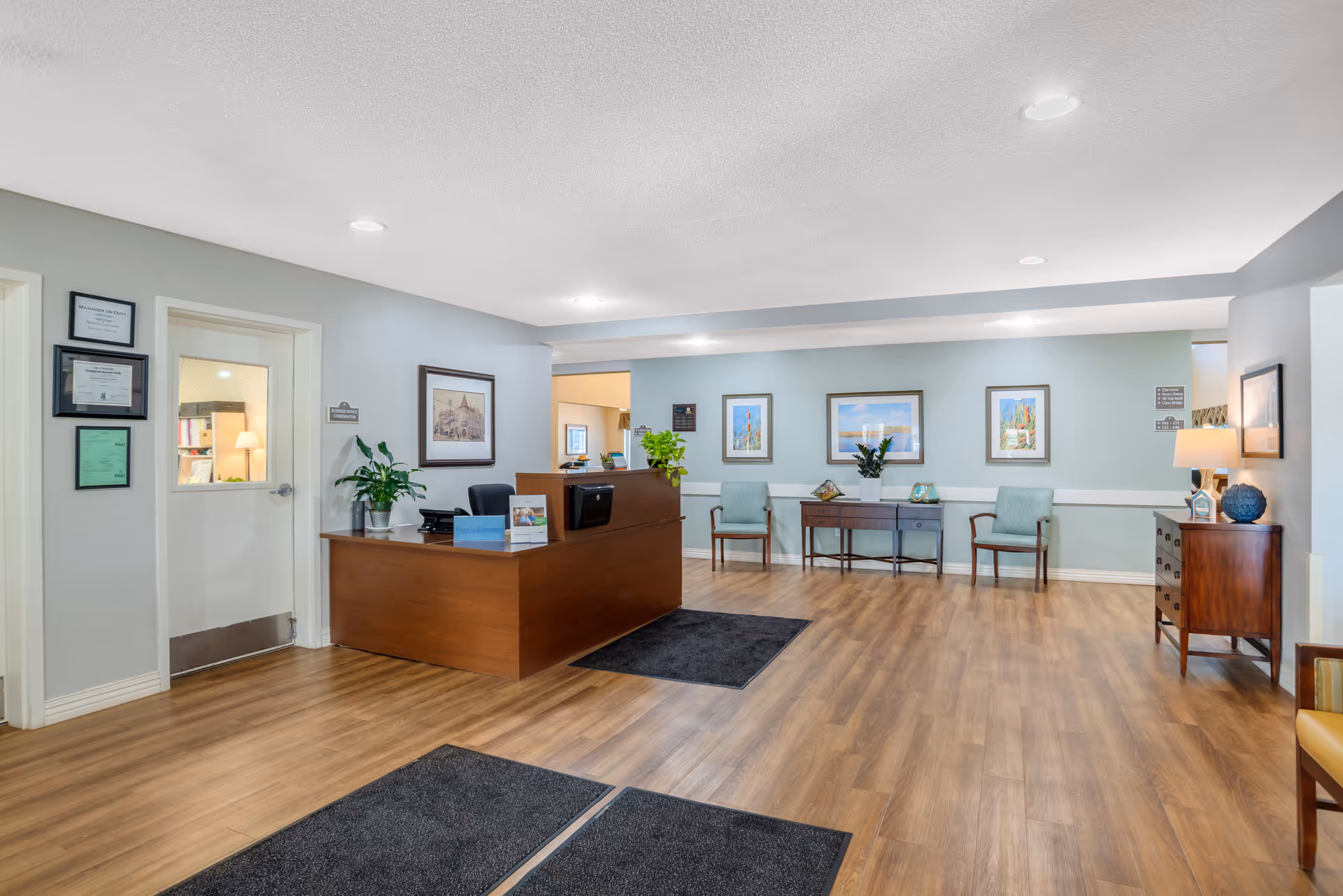 Reception area of a senior living facility with a wooden desk, office chair, potted plants, framed pictures on the walls, and seating area with chairs and a small table. The floor is wood with black mats near the entrance, and the walls are painted light blue and gray.