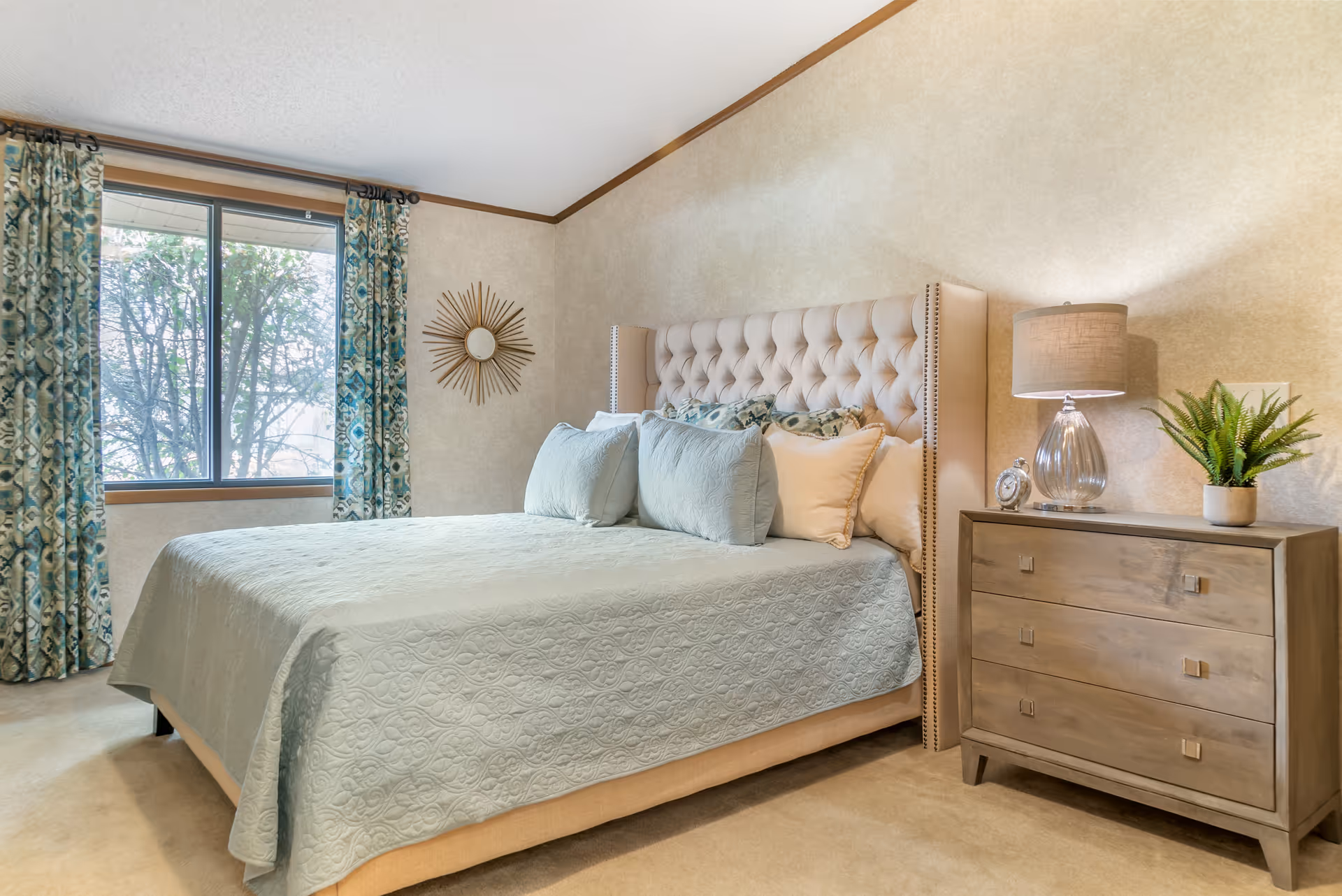A cozy bedroom with a large tufted beige headboard bed covered in a light blue quilt and multiple pillows. Next to the bed is a wooden nightstand with three drawers, a glass lamp, a small clock, and a potted plant. A window with patterned blue and beige curtains allows natural light to enter the room. A decorative sunburst mirror hangs on the wall beside the window.