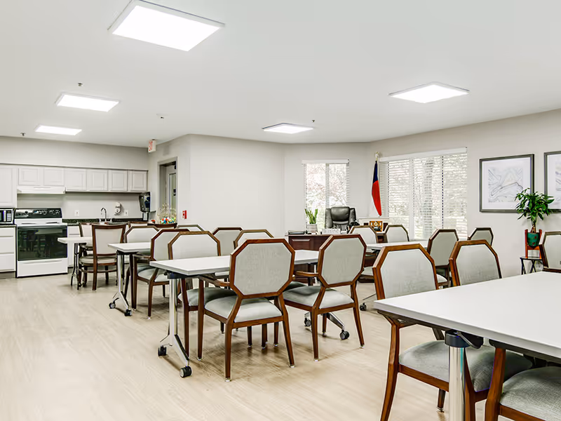 A bright and clean common area with multiple tables and chairs arranged neatly. In the background, there is a kitchen area with white cabinets, a stove, microwave, and refrigerator. Large windows let in natural light, and there is a desk with a chair near the windows. The room has light-colored flooring and white walls, with some framed artwork and a potted plant.