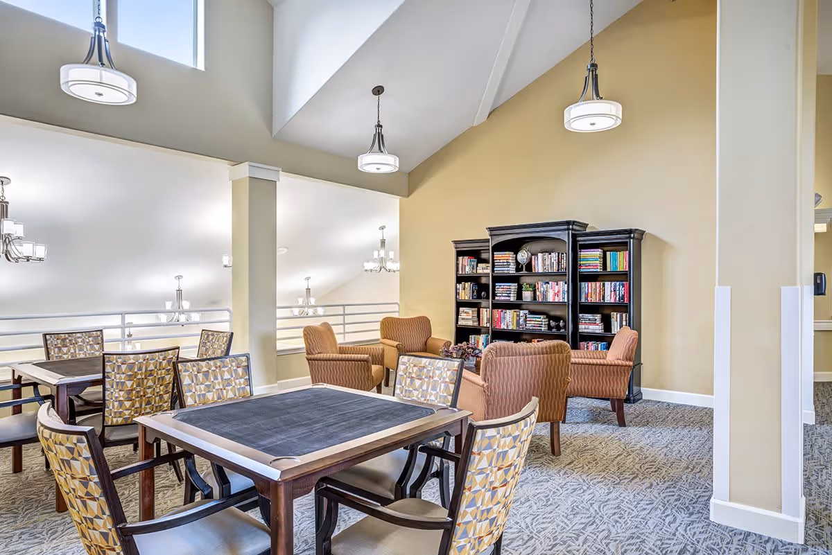 A bright and spacious senior living common area with high ceilings and hanging light fixtures. The room features several tables with patterned chairs and a seating area with four upholstered armchairs arranged around a small table. A large black bookshelf filled with books and decorative items is against a beige wall.