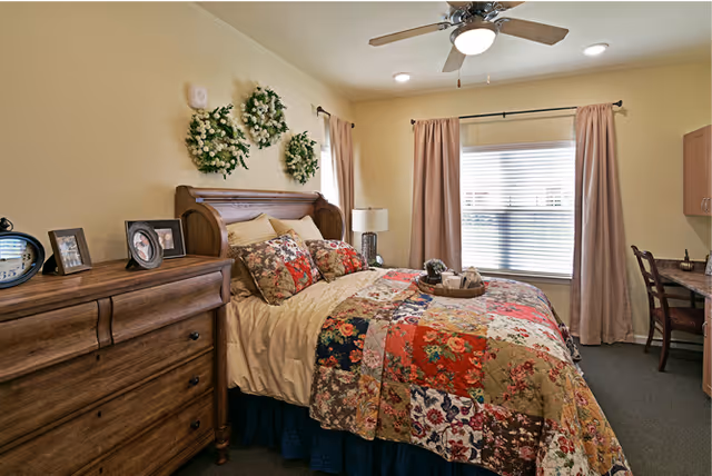 A cozy bedroom with a wooden bed frame and a colorful patchwork quilt on the bed. There are three decorative wreaths hanging on the wall above the bed. A wooden dresser with framed photos and a clock is on the left side. A window with beige curtains lets in natural light. A small desk and chair are positioned against the right wall.
