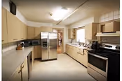 A spacious kitchen with beige cabinets, a stainless steel refrigerator, a microwave, a coffee maker, and a stove with an oven. The kitchen has a tiled floor and a window above the sink letting in natural light.