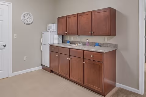 A small kitchenette area with wooden cabinets, a countertop with a sink, a white refrigerator, a microwave, a paper towel holder, and a wall clock. The walls are painted beige and the floor is carpeted.