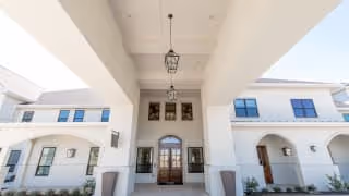 Front entrance of a white Mediterranean-style building with a covered portico, hanging lanterns, and double wooden doors.