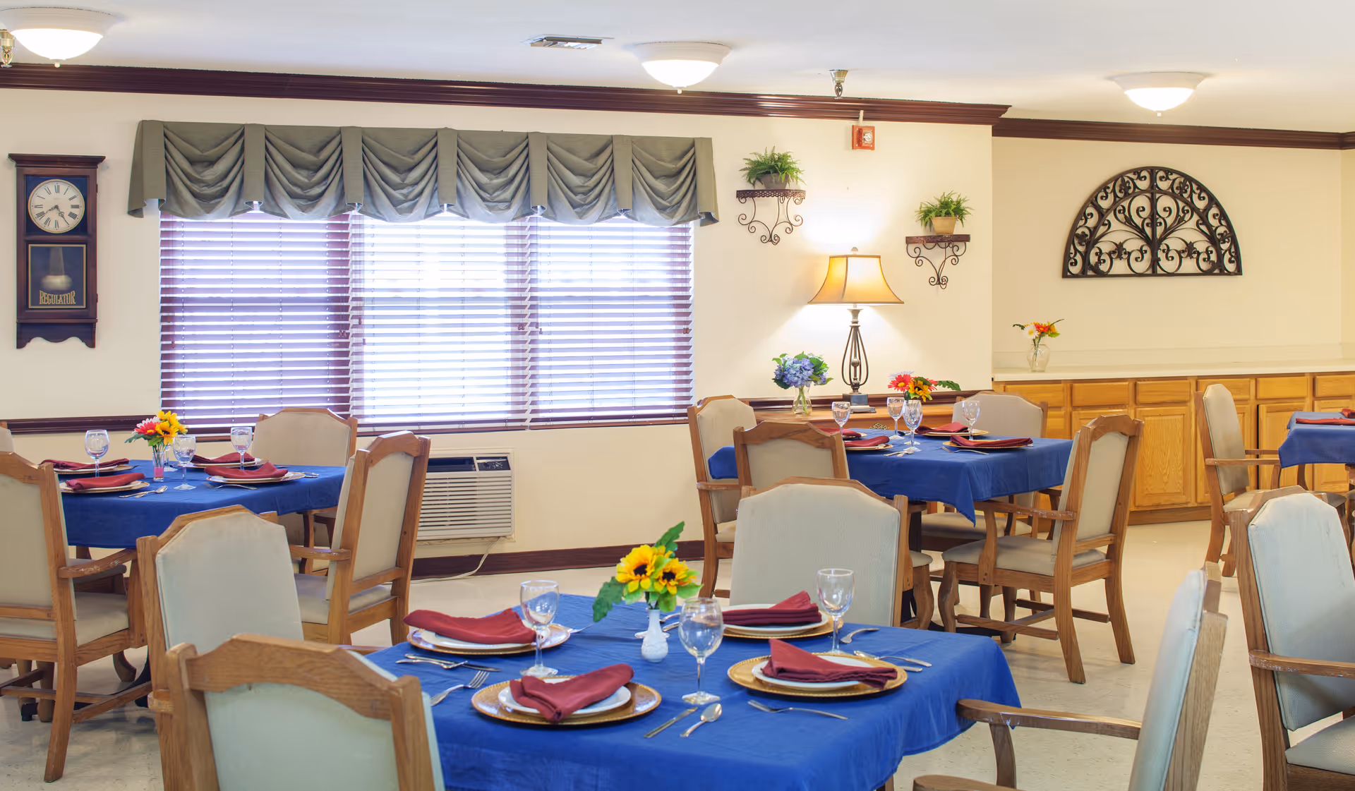 A dining room with several round tables covered with blue tablecloths, each set with plates, silverware, wine glasses, and red napkins. The room has wooden chairs with light-colored cushions, large windows with blinds and valances, a wall clock, decorative plants, a table lamp, and a decorative wrought iron wall piece.