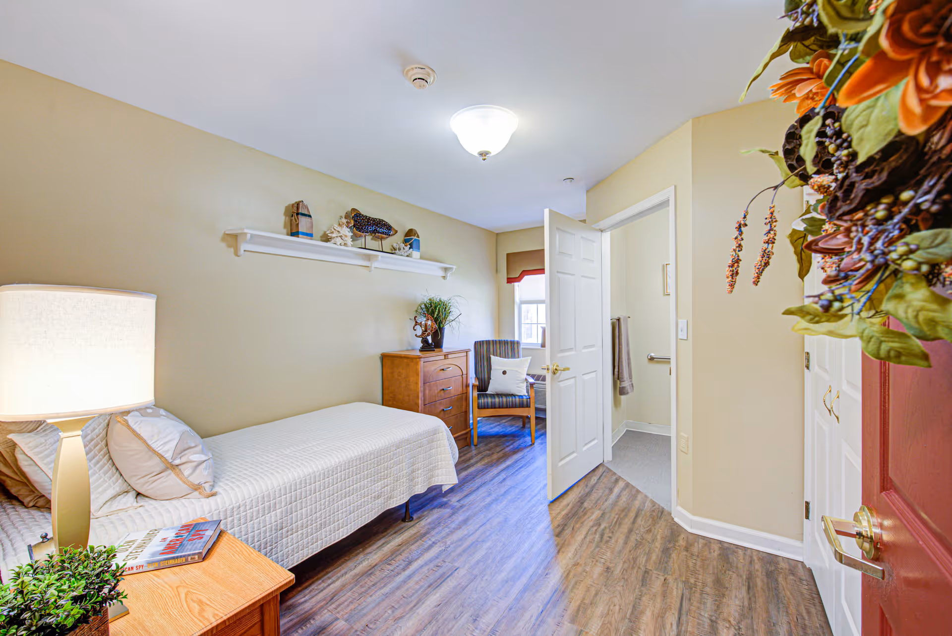 A small, cozy bedroom with a single bed covered in a white quilt, a wooden nightstand with a lamp and a book, a wooden dresser with decorative items on top, a striped armchair with a pillow near a window, and an open door leading to a bathroom. The floor is wood laminate, and the walls are painted light beige. There is a decorative wreath on the partially visible red door on the right.