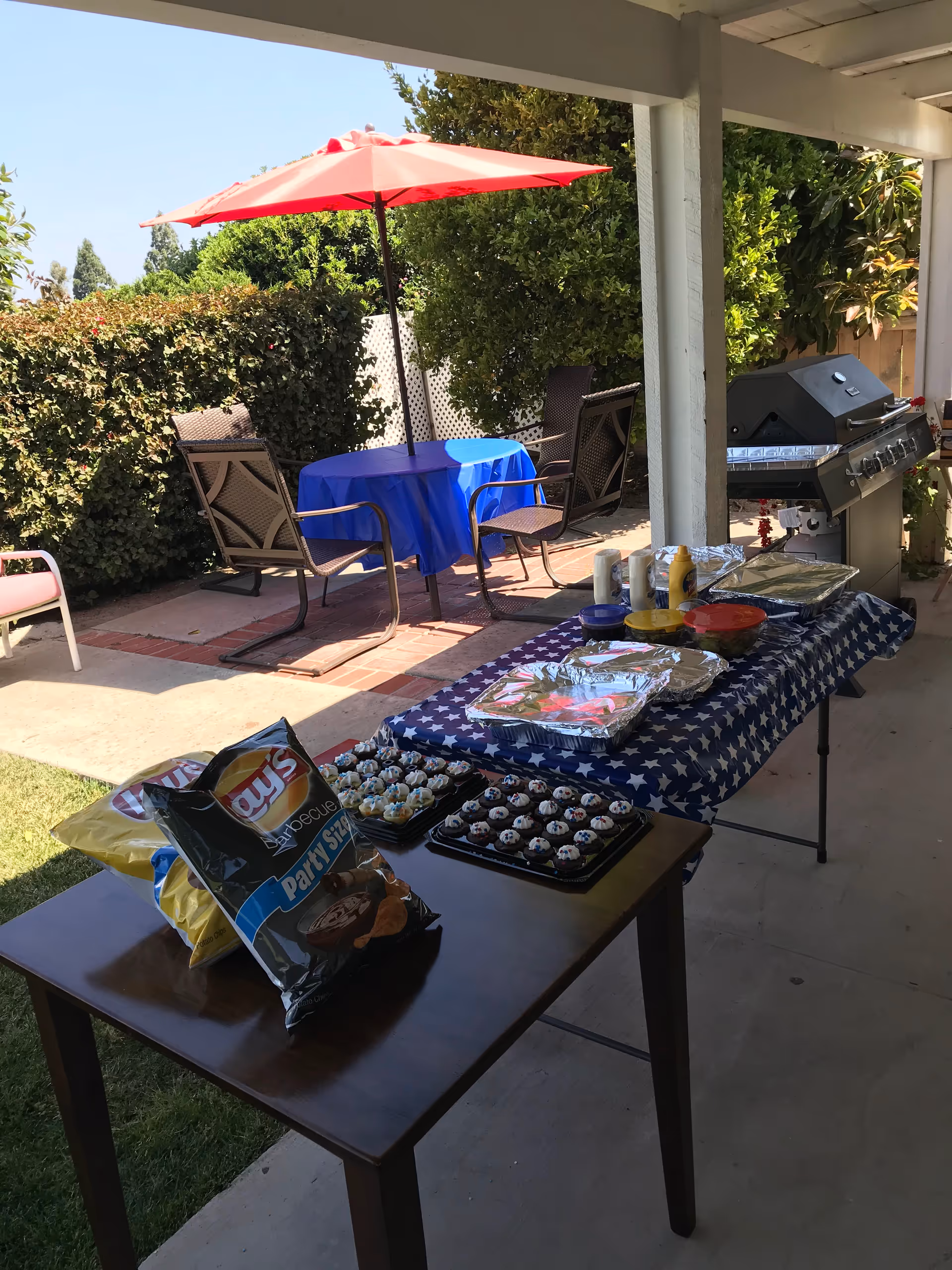 Outdoor patio area with a table covered in a star-spangled tablecloth holding trays of food, condiments, and bags of chips. Nearby is a barbecue grill and a round table with a blue tablecloth and a red umbrella, surrounded by chairs. Green bushes and trees are visible in the background under a clear sky.