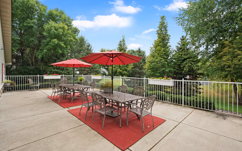 Outdoor patio area with two metal tables and chairs set on red rugs. Each table has a large red umbrella providing shade. The patio is surrounded by a white metal railing and overlooks a green lawn with trees and shrubs under a partly cloudy blue sky.