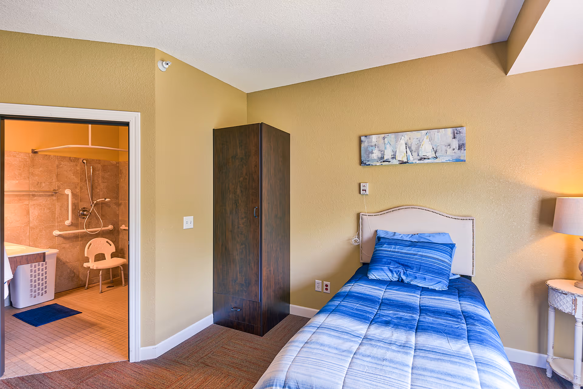 A single-bed bedroom with blue bedding, a wardrobe and nightstand, and an open door showing an accessible bathroom with grab bars and a shower chair.