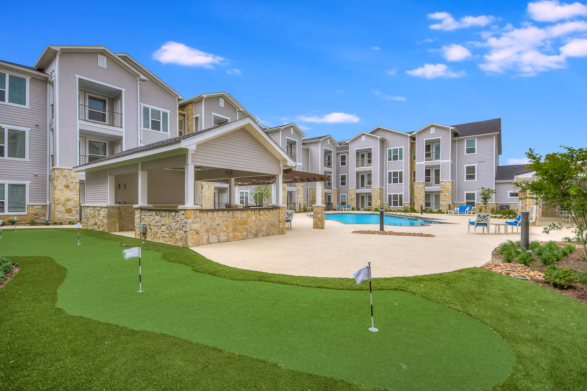 Outdoor area of a senior living facility featuring a putting green with small flags, a covered pavilion with stone pillars, a swimming pool surrounded by lounge chairs, and a multi-story residential building in the background under a blue sky with scattered clouds.