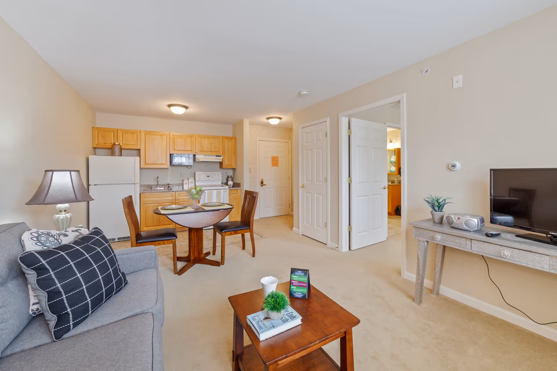 A bright and cozy senior living apartment featuring a small kitchen with wooden cabinets, a white refrigerator, stove, and microwave. In front of the kitchen is a round dining table set with two chairs. To the left, there is a gray sofa with patterned pillows and a lamp on a side table. On the right side, a wooden console table holds a small plant, a radio, and a TV. The room has beige walls and carpet, with two open doors leading to other rooms.