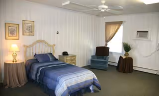 A simple bedroom with a wooden bed, nightstand, lamp, blue bedding, and chairs.