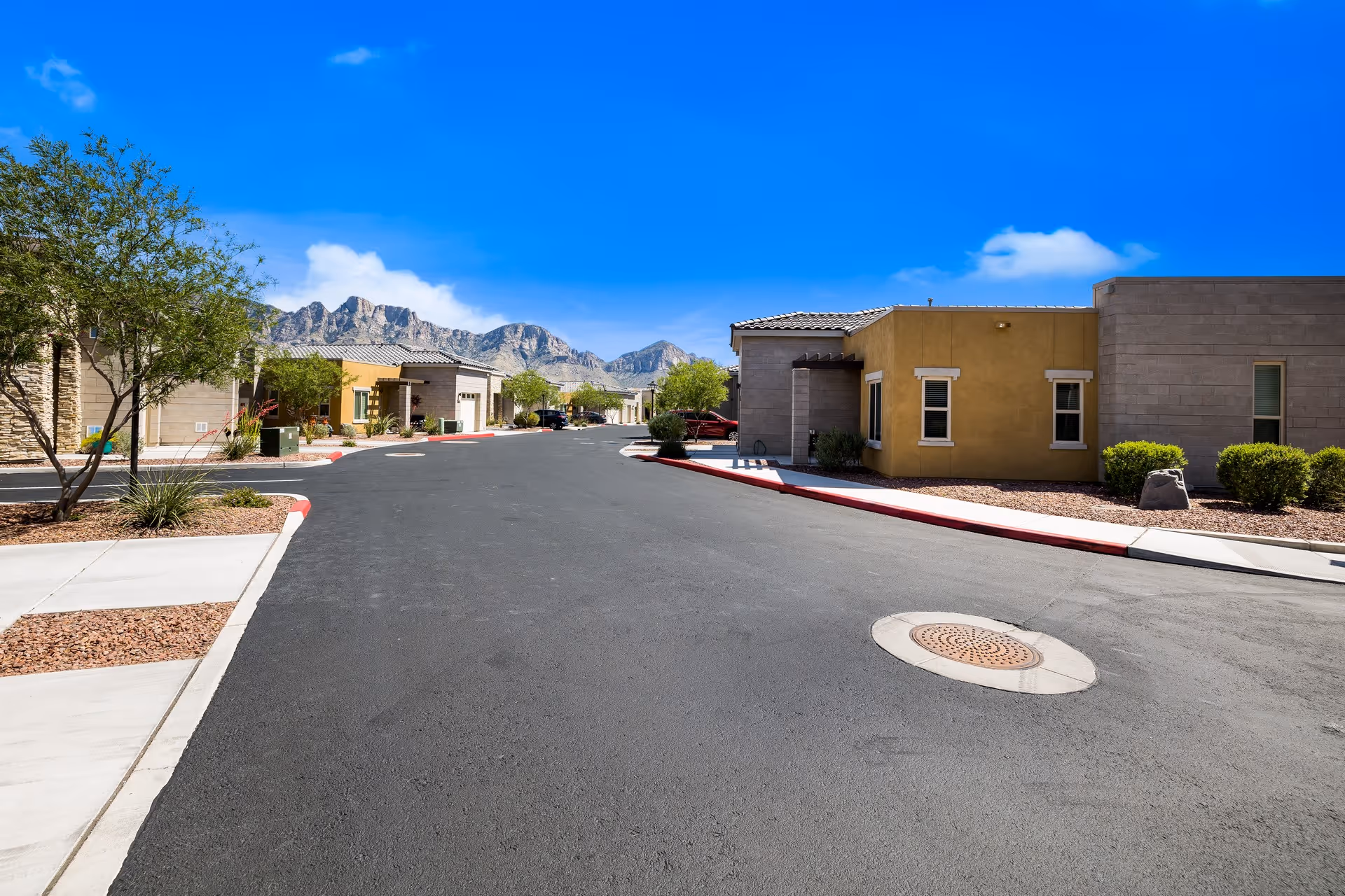 A paved road within a senior living facility with single-story buildings on either side, desert landscaping with small trees and bushes, and mountains visible in the background under a clear blue sky.