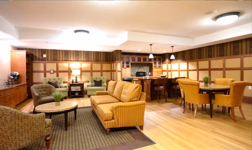 A cozy common area in a senior living facility featuring a yellow striped sofa, several armchairs, a round table with four yellow upholstered chairs, and a kitchen area with bar stools in the background. The room has wooden paneling on the walls, warm lighting, and a wooden floor.