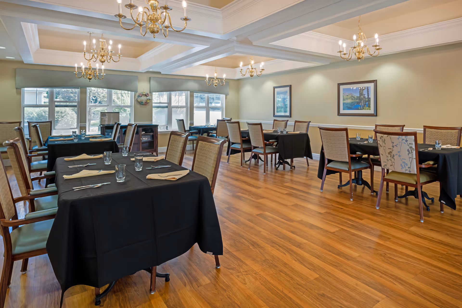 Spacious dining room with multiple tables covered in black tablecloths, chairs, chandeliers, and large windows.