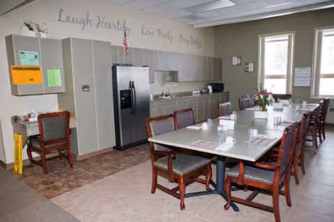 Communal dining room with long tables, chairs, and a kitchenette in a senior facility.