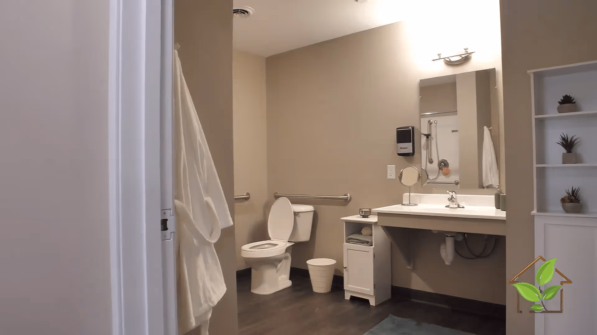 A clean and accessible bathroom with a white toilet, a white sink with a mirror above it, a small white cabinet with towels, a white waste bin, and a white robe hanging on the wall. There are grab bars near the toilet and a shower area visible in the mirror reflection. The walls are beige and the floor is dark wood. A white shelving unit with small potted plants is on the right side.